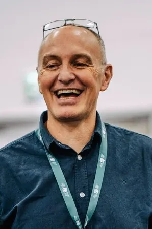 A man wearing a blue t-shirt and a lanyard. He is smiling a lot and has glasses perched on his head.