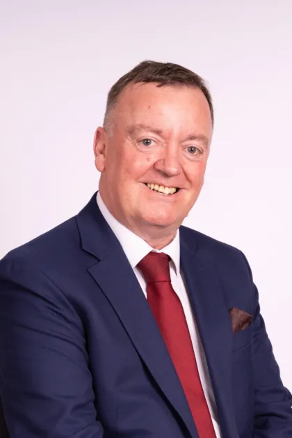 A white man with short brown hair. He is smartly dressed in a blue jacket and red tie.