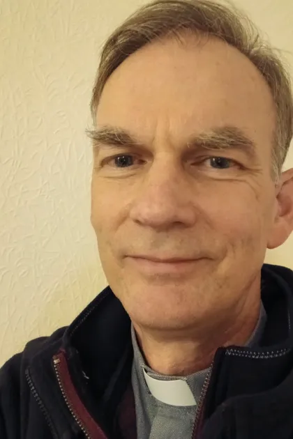 A white man with short mousy hair. He is wearing a white dog collar and grey shirt, with a dark jacket.