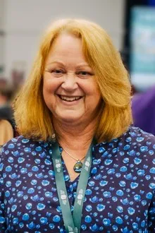 A woman with strawberry blonde shoulder-length hair, wearing a blue dress. She looks relaxed and is smiling.